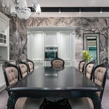 Black table with chairs placed near white cupboards and modern appliances in stylish spacious kitchen with mirror and creative wallpaper